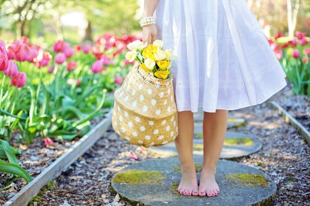 A woman in a white dress carries a basket of yellow flowers while walking barefoot in a tulip garden.